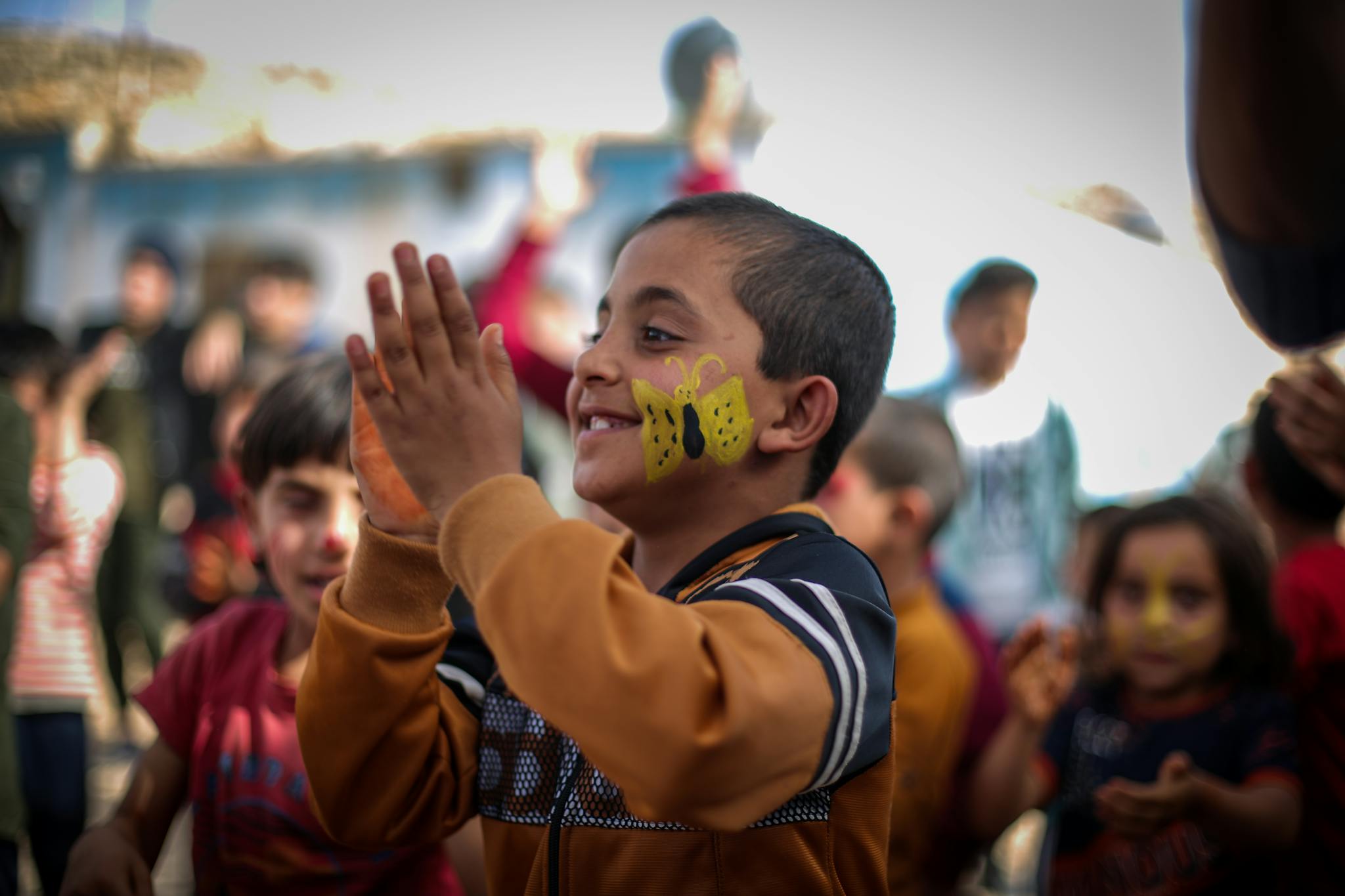 A smiling child with butterfly face paint claps joyfully among friends in Idlib, Syria.